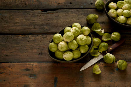 From above of fresh green Brussels sprouts in bowls placed on rustic wooden table with knife