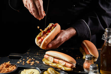 Chef preparing a tasty hot dog with a frankfurter on a fresh roll adding extra trimmings sprinkling crispy bacon bits onto the pickles and sausageの写真素材