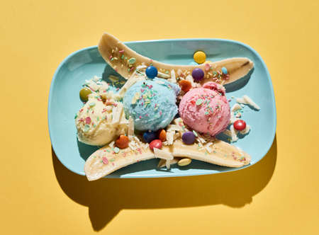 Top view of delicious banana split dessert with colorful ice cream scoops decorated with dragee and sprinkles served on blue plate on yellow backgroundの写真素材