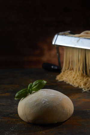Raw pastry with basil leaves placed on wooden table against metal machine for making spaghetti in dark studioの写真素材
