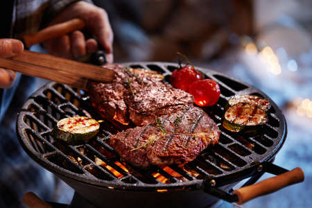 High angle of crop male cook turning appetizing beef steak and vegetables on BBQ grid in winter eveningの写真素材