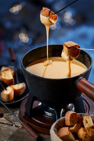 From above of crispy bread croutons being dipped in liquid cheese fondue melted in pot on gas burner in winterの写真素材