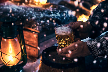 Crop faceless male with glass jar of cold lager beer sitting in winter yard at snowy night in winterの写真素材