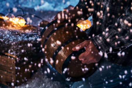 High angle of crop unrecognizable male with wooden barrel of brewed beer standing in winter garden at nightの写真素材