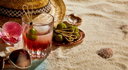 High angle of glass of refreshing drink with ice cube and olive near straw hat sunglasses on sandy beach in sunny resortの写真素材