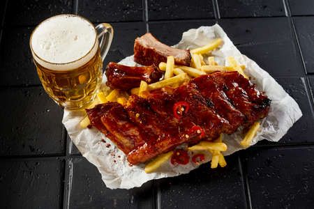 High angle of delicious grilled spicy ribs served with fries placed near jar of beer with foam on black tableの写真素材