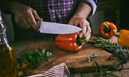 Crop unrecognizable male chef in checkered shirt cutting fresh bell peppers on wooden chopping board while standing at table with olive oil bottle and rosemary sprigs during cooking in kitchenの写真素材