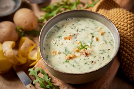 From above of appetizing homemade cream rocket soup with croutons served on wooden cutting board near peeled potatoes in kitchenの写真素材