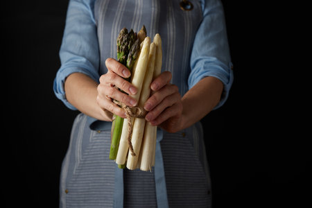 Crop unrecognizable female cook in apron holding bunch of raw healthy white and green asparagus against black background in studioの写真素材