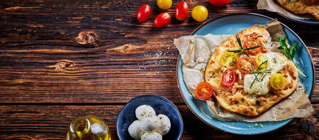 Top view of delicious homemade crispy flatbread pizza with cherry tomatoes mozzarella and herbs served on plate near ingredient on wooden tableの写真素材