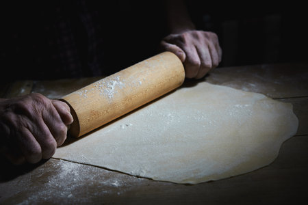 Crop anonymous male chef rolling out dough on wooden table covered with flour in bakery