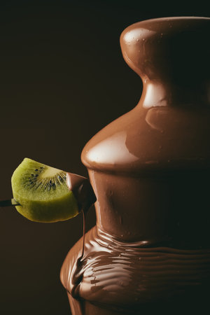 Unrecognizable person dipping piece of sweet kiwi into tasty chocolate fountain during party against black backgroundの写真素材