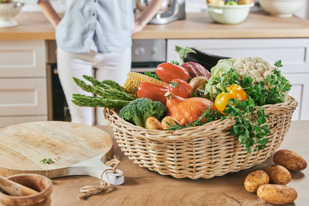 Fresh assorted vegetables in wicker basket near chopping board placed on wooden counter with crop woman in backgroundの写真素材