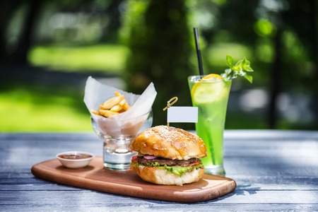 Perfect meat burger with french fries and fresh cold lemonade. On the cutting board, and green summer background.の写真素材