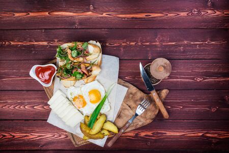 Big breakfast. Bruschettas, fried eggs, fried potatoes and cheese with sauce. There is a black pepper and cutlery. Top view on a wooden background.の写真素材