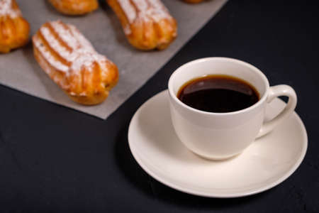 Black coffee in a white cup and eclairs with cream on a dark background