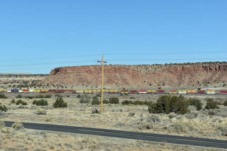 McCartys, New Mexico, USA - November 21, 2021: Cargo Train Travels Through the Desert, Next to a Road, with Mountains in the Background and Electricity Wires in the Foregroundのeditorial素材