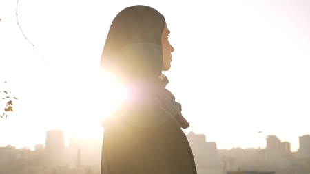 Closeup profile shoot of young attractive female silhouette in hijab looking forward being turned to the side with urban city on the backgroundの写真素材