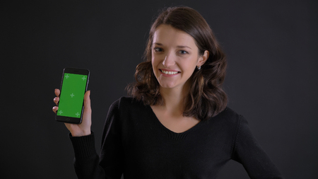 Portrait of cute young brunette woman showing green screen of smartphone joyfully on black background.の写真素材