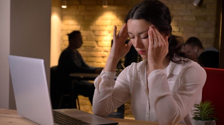 Closeup shoot of young pretty caucasian businesswoman having a headache while typing on the laptop indoors on the workplaceの写真素材