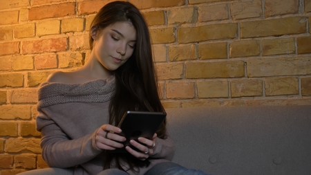 Closeup portrait of young attractive caucasian female texting on the tablet while sitting on the couch indoors in a cozy apartmentの写真素材