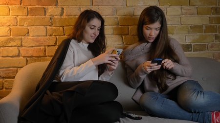 Closeup portrait of two young pretty caucasian girls using the phones while resting on the couch indoorsの写真素材