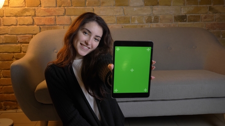 Closeup portrait of young pretty caucasian female browsing on the tablet and showing green chroma screen to camera in a cozy home indoorsの写真素材