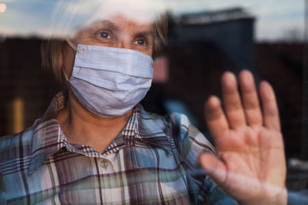 Elderly caucasian woman wearing hand made protective face mask, in nursing care home, looking outside window with sadness in her eyes, self isolation due to the global COVID-19 Coronavirus pandemicの写真素材