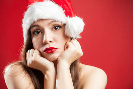 Worried depressed attractive caucasian brunette woman dressed as Mrs Santa Claus,wearing red white hat,chin on hands,isolated on red background,copy space,New Year 2021 COVID-19 Coronavirus pandemicの写真素材