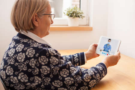 Retired senior elderly woman talking to GP physician via virtual telemedicine video call,holding...