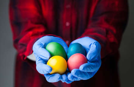 Person wearing blue medical protective gloves holding colored Easter eggs, holiday amid global...