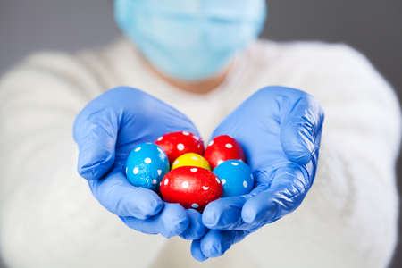 Person wearing face mask and blue protective medical gloves holding chocolate Easter eggs, festive...