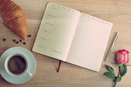 red rose and a Cup of coffee with a croissant and diary with days and month of the week in Russian and English languages on a wooden table morning Breakfast of planingの写真素材