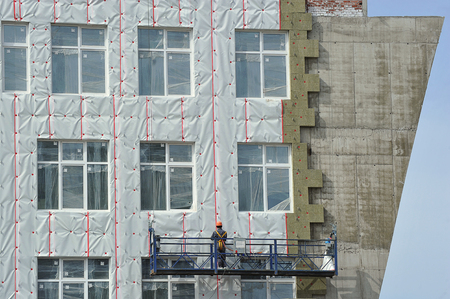 workman in a hanging cradle at the construction site insulates the walls of the house insulationの写真素材