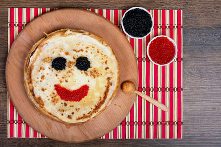 Round pancakes with a face made of red and black caviar, lie on the board. Close-up, top view, Flat Lay, Copy space, place for text. Shrovetide, East Slavic traditional holiday of Ukraine.の写真素材