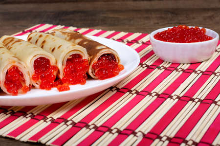 A plate with fried pancakes, twisted in a straw and stuffed with red salmon caviar, stands on a bamboo red and white stand. Close-up side view. Shrovetide, East Slavic traditional holiday of Ukraine.の写真素材