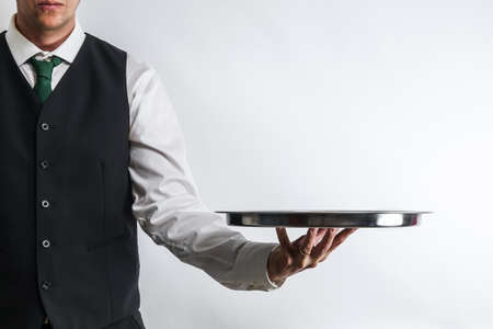 Butler / waiter in white shirt and black suit vest carrying an empty silver tray. Copy space for text on white background.の写真素材