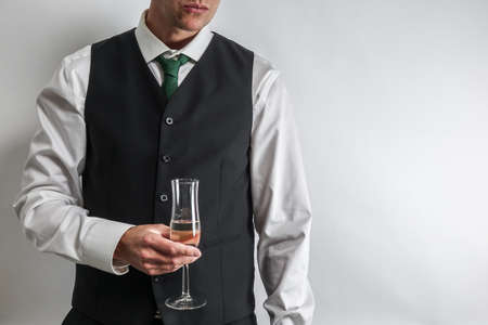 Well dressed man in white shirt and black suit vest holding a glass of champagne, toast / cheering. White background with copy space for text.の写真素材