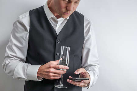 Well dressed man in white shirt and black suit vest holding a glass of champagne and texting with...