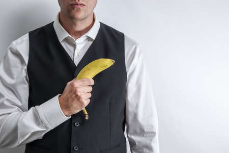 Well dressed man in white shirt and black suit vest holding a banana like a gun. Concept of fake gangster / radical militant vegan. White background with copy space for text.の写真素材