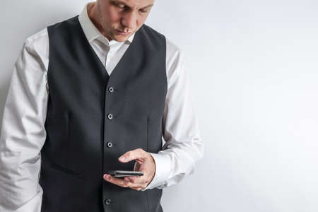 Well dressed man in white shirt and black suit vest loooking at his smart phone. White background with copy space for text.の写真素材
