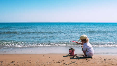 A child plays with a pink plastic bucket and shovel in the sand on a beach, close to the sea. Crete, Greece.の写真素材