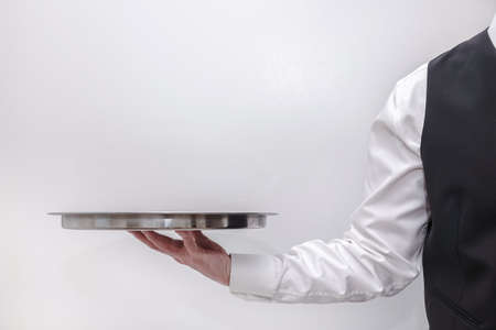 A waiter / butler in white shirt and black suit vest carrying an empty silver tray. Copy space on white background.の写真素材