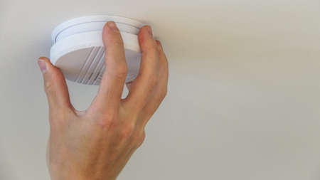 A person replaces batteries in a automatic smoke detector fire alarm in the ceiling. - Image