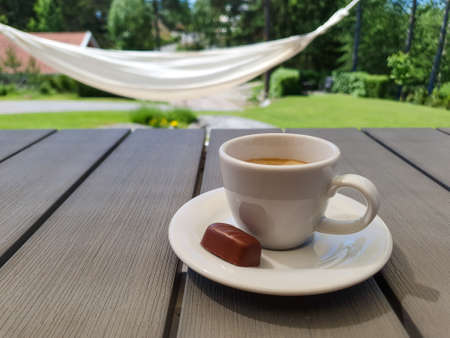 Cup of espresso with small chocolate praline on gray table with hammock and green sunny garden in the background.の写真素材