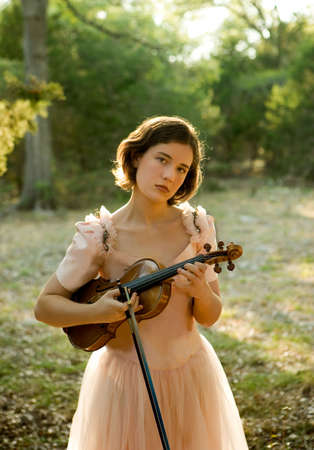 A pretty young woman/teenager in a vintage dress holding a violin and bow in a woodland setting. Subject is backlit, and photo has a nostalgic feel. Plenty of background at top of picture for type, etc.の写真素材
