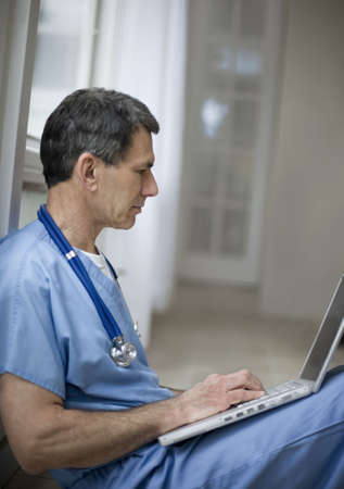 Middle-aged male doctor or surgeon sitting on floor with his laptop on a break, wearing blue scrubs