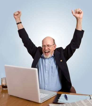 Senior man seated at desk, looking at computer, cheering over good news he has just received.の写真素材