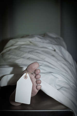 Dark atmospheric photo of body covered with a sheet in a morgue, feet toward camera with blank tag...