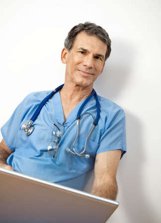 Mature doctor sitting on floor, leaning against wall, in blue scrubs, working on his laptopの写真素材